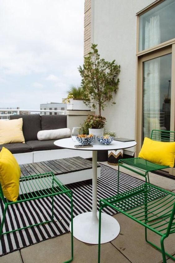 a Scandinavian balcony with a sofa and side table, a striped rug, a coffee table, green chairs and yellow pillows and some plants