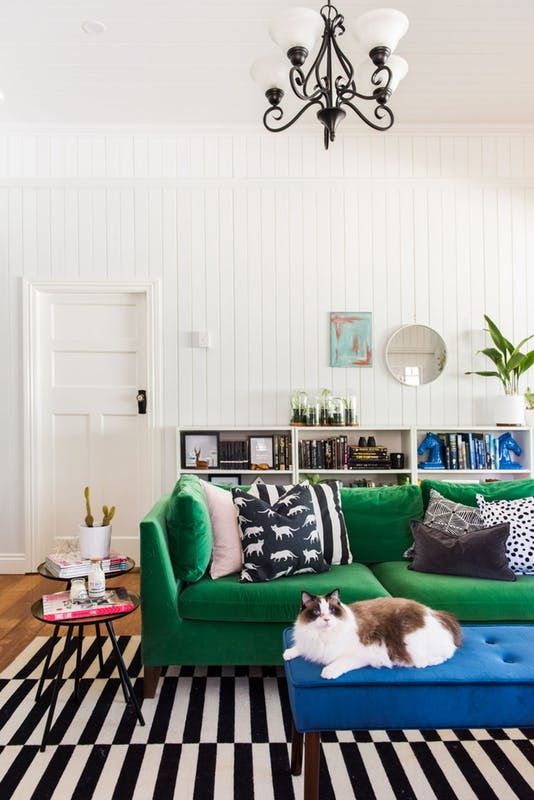 a Scandinavian living room with a striped rug, a green sofa with printed pillows, a bold blue ottoman, a shelving unit with decor