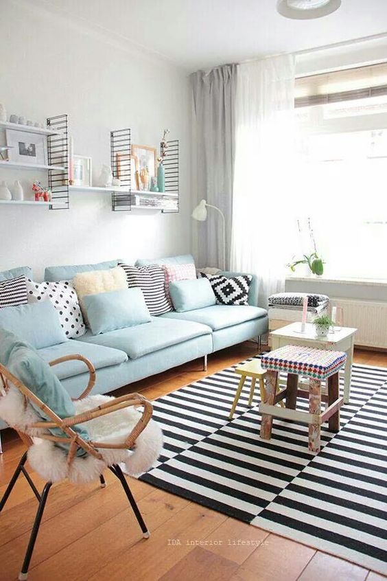 a Scandinavian living room with an aqua sofa with pillows, stools, a rattan chair, a stripe rug and some wall-mounted shelves