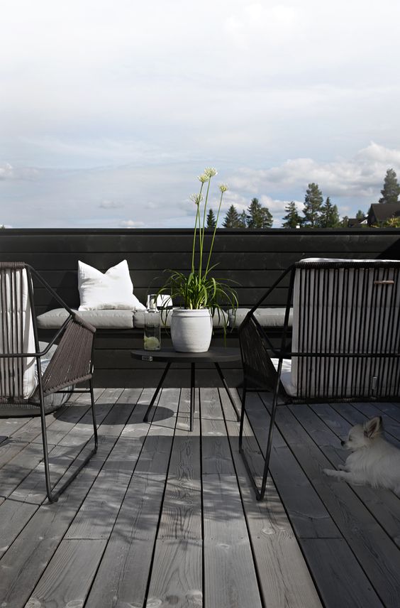 A Scandinavian terrace with a black fence, a built in sofa and black chairs, a coffee table and some blooms and greenery