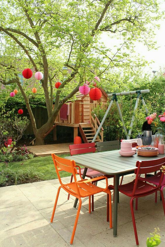 a backyard done with a green dining table, orange and red chairs, a tree decorated with pink and red paper lamps is fun