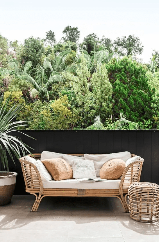 a balcony with a rattan daybed, a side table and a potted plant plus lots of pillows to style the sofa