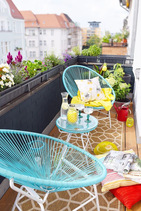 a balcony with bright utrquoise chairs and a side table, uellow blankets and colorful pillows, greenery and blooms in pots