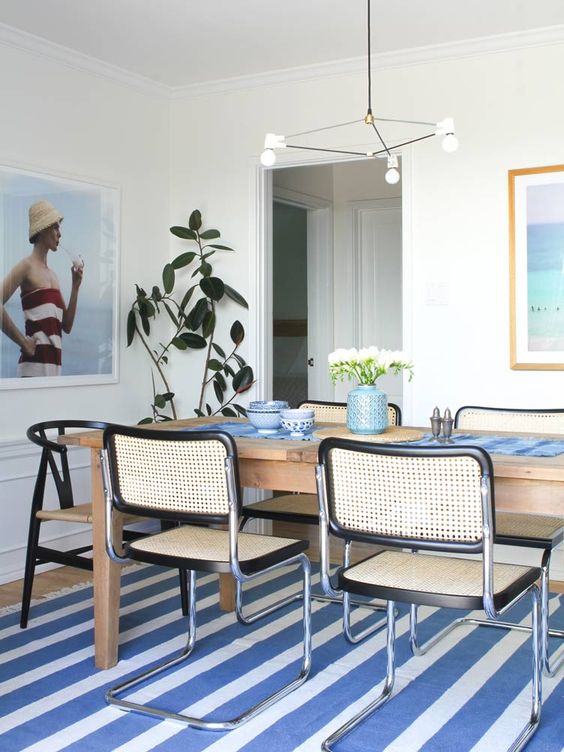 a beach dining room with a striped blue rug, a table, cane chairs, a potted plant and an artwork and a modern chandelier