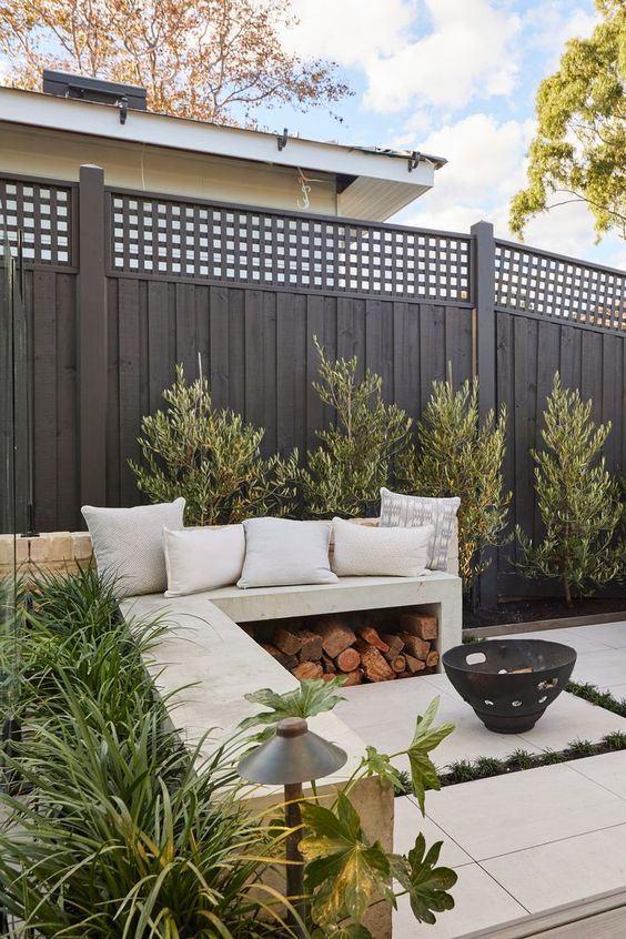 a black fence, a concrete built-in bench with firewood storage, greenery around and a small fire pit