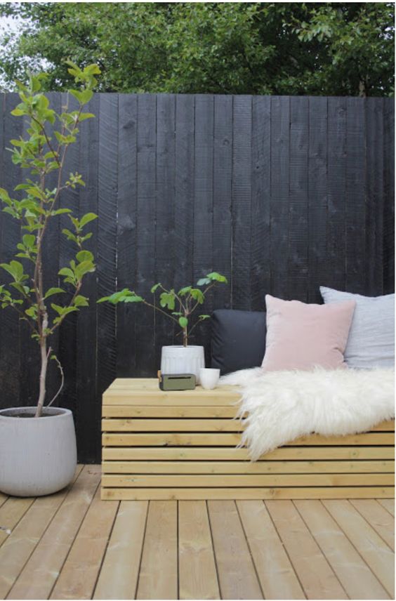 a black fence, a light-stained bench with pastel pillows, potted greenery and a light-stained deck to make the space fresh