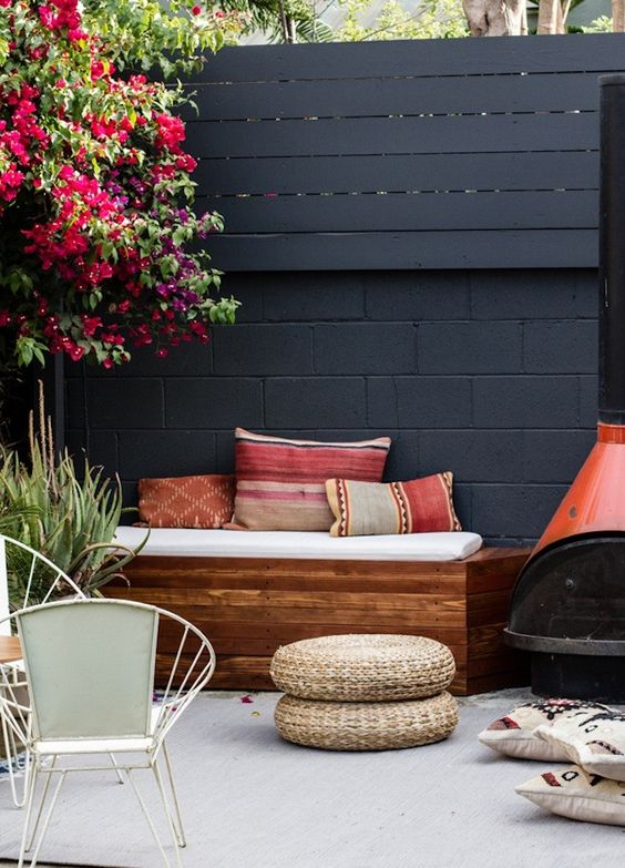 a black fence, a stained seat with pillows, a red hearth, some blooms and greenery, white chairs and jute poufs