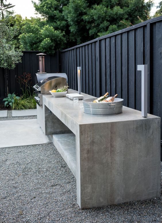 a black fence and a concrete outdoor kitchen along it, with garden beds with greenery in the corner