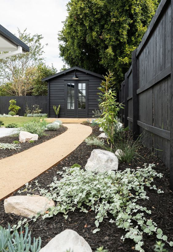 a black fence lined up with a garden bed with rocks, greenery and grasses to make the fence look fresh and cool