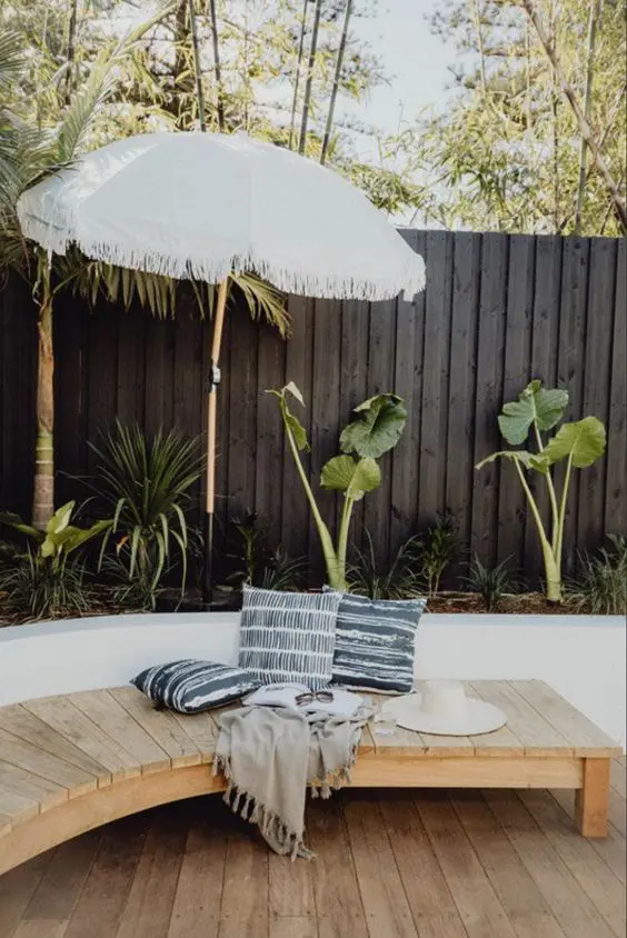 a black fence lined up with a raised garden bed with tropical plants, a curved bench, an umbrella and some printed textiles