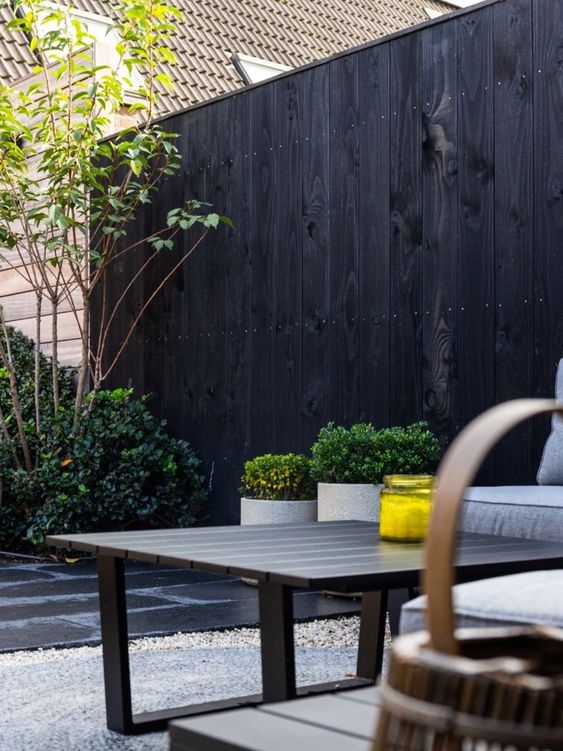 A black fence with greenery and plants in pots along it, neutral furniture, a dark stained coffee table and a candle