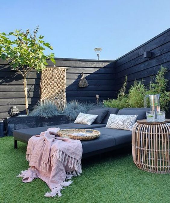 a black fence with grey loungers, a raised garden bed with greenery and some boho decor, a side table and some accessories
