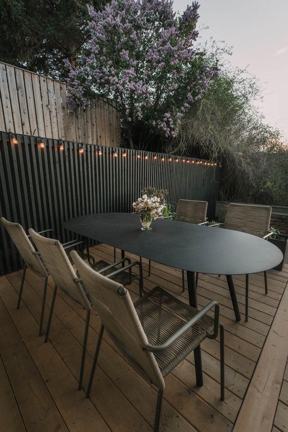 a black fence with lights, a black curved table, woven chairs, some greenery are a cool and bold modern space