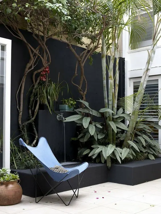 a black fence with trees and greenery in a raised garden bed, a blue butterfly chair and some blooms are a cool modern combo