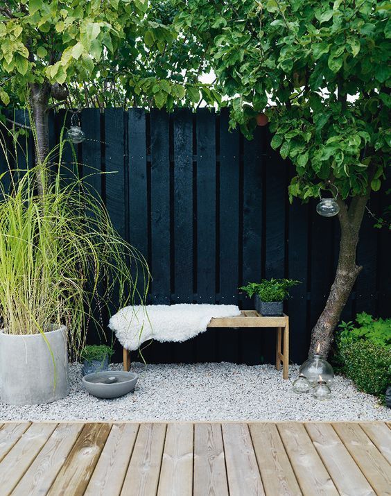 a black wooden fence, a bench with a rug and greenery, greenery and a tree are a stylish and cool combo for a modern space