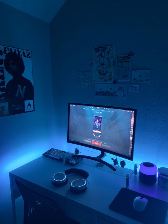 A blue gaming nook with a desk and a chair, a PC and blue built in lights, posters and a mini gallery wall