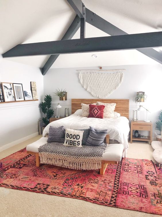 a boho bedroom with a bold boho rug, a bed with neutral bedding, a bench with pillows and some boho decor