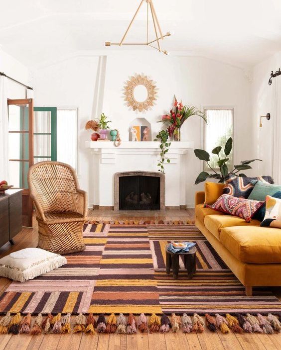 a boho living room with a colorful striped rug, a yellow sofa with printed pillows, rattan chair, a fireplace with some pretty decor