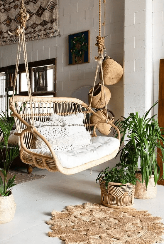 a boho porch with a double rattan chair with neutral cushions and pillows, a jute rug, some baskets hanging and potted plants