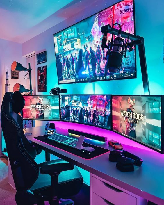 a bold and colorful gaming space with a large desk, some screens, a chair, a lamp and some posters on the wall plus blue and purple neon lights