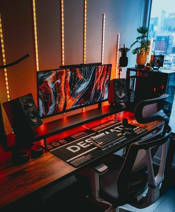 a bold and moody gaming nook with a desk, a screen, a PC, a black chair and strip lights on the wall plus greenery