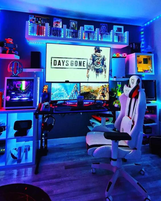 a bold blue gaming space with a PC and several screens, a chair, some devices, shelves with books and decor and some built-in lights