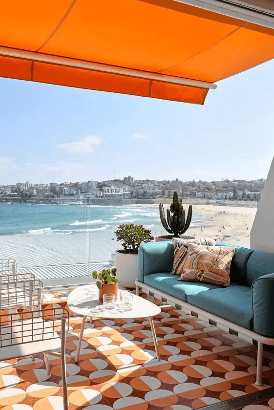 A bright beach terrace with an orange rug and a roof over it, a turquoise sofa and comfy chairs, a mid century modern table