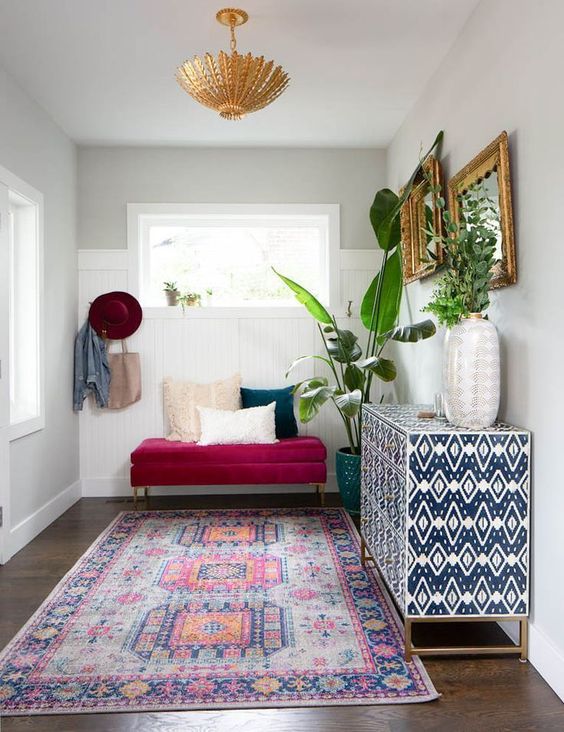 a bright boho entryway with a boho rug, a fuchsia bench with pillows, a printed sideboard, mirrors and a gold chandelier