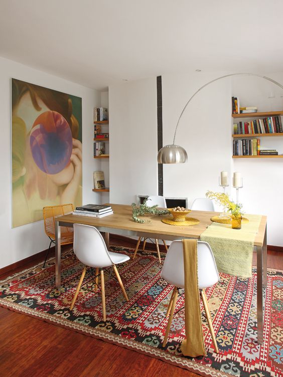 a bright dining room with a bold artwork, a bold boho rug, a stained table and mismatching chair, built-in bookshelves and a floor lamp