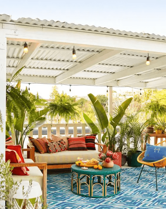 a bright terrace with a blue printed rug, neutral furniture and colorful pillows, an emerald table and potted plants all around