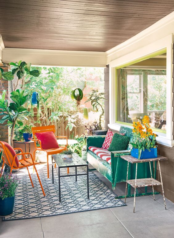 a bright terrace with a green sofa and colorful pillows, orange chairs, a green table and potted plants and flowers