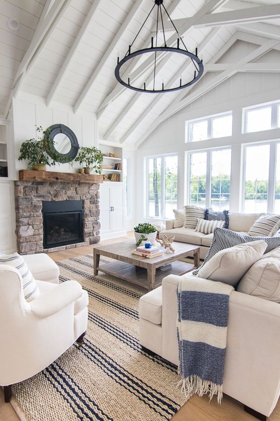 a coastal farmhouse living room with a stone fireplace, a striped rug, white seating furniture, a coffee table and a heavy chandelier