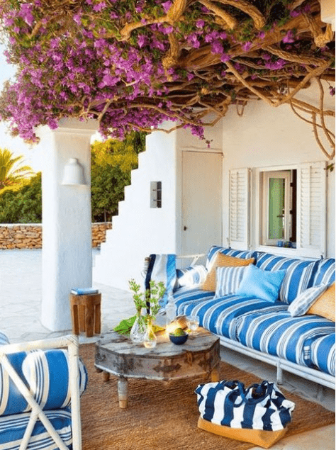 a colorful Mediterranean terrace with bright striped furniture, bright blooms over the terrace, a vintage table and a jute rug