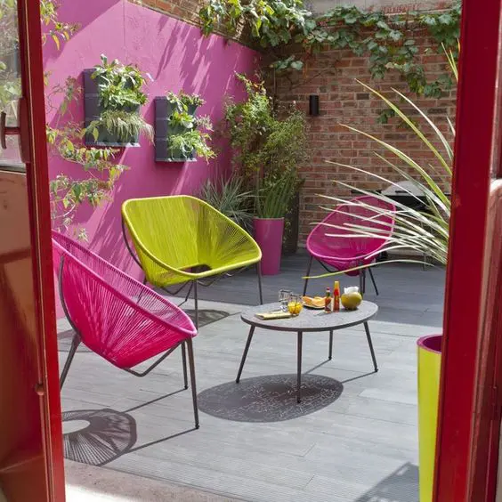 a colorful modern terrace with a fuchsia wall, fuchsia chairs and a lemon yellow loveseat, a coffee table and some greenery