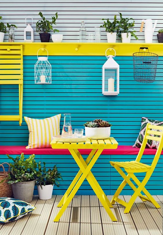 a colorful outdoor space with a blue and yellow wall and a built-in seat with a pink cushion, a yellow folding table and chair and some potted greenery