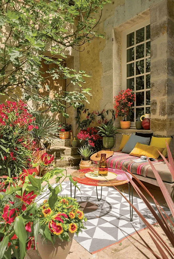 a colorful summer terrace with a daybed with colorful pillows, a woven coffee table, a printed rug, potted blooms and plants feels Mediterranean
