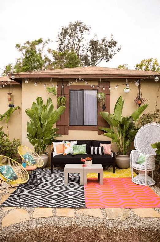 a colorful summer terrace with bright printed rugs, a black sofa, a peacock chair, yellow round chairs, printed pillows and potted tropical plants