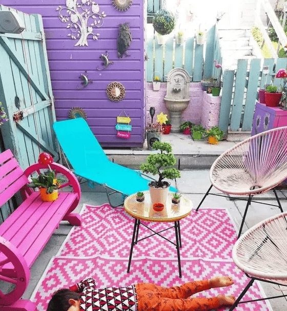 a colorful terrace with a purple wall, a hot pink bench, a bold rug, a turquoise lounger and some bright decor