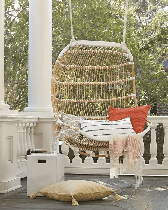 a double hanging rattan chair with printed pillows and a pillow on the floor, a small side table compose a cool porch for relaxing