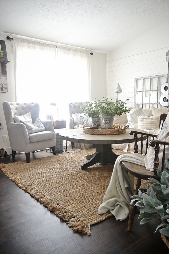 a farmhouse living room with white seating furniture, a round stained table, a jute rug, a wooden rocker chair, potted greenery and some chic decor