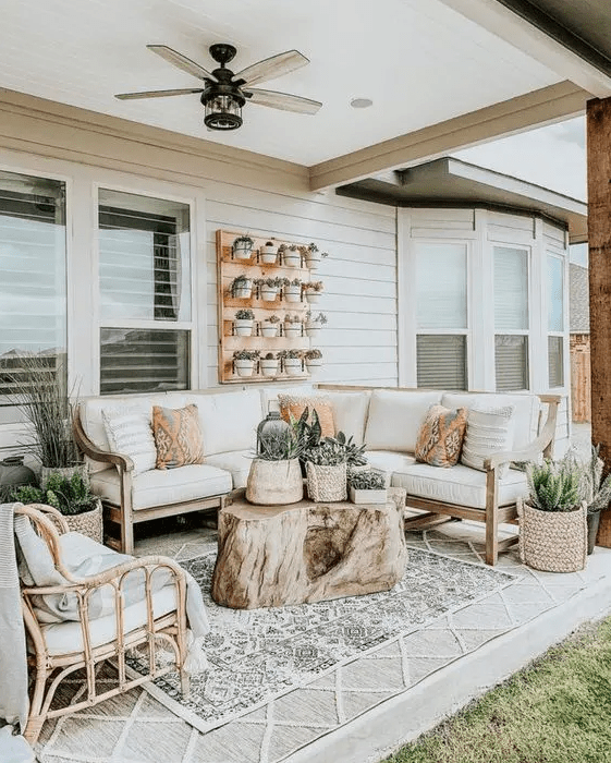 a farmhouse porch with a wooden corner sofa, a rattan chair, printed pillows, blankets and layered rugs and some potted greenery