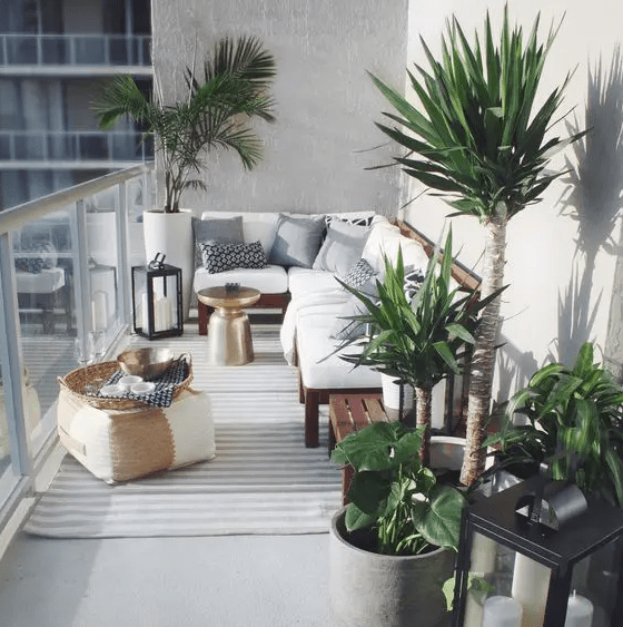 A lovely and welcoming balcony with a rich stained wooden bench with white upholstery, potted plants, a small pouf and a striped rug