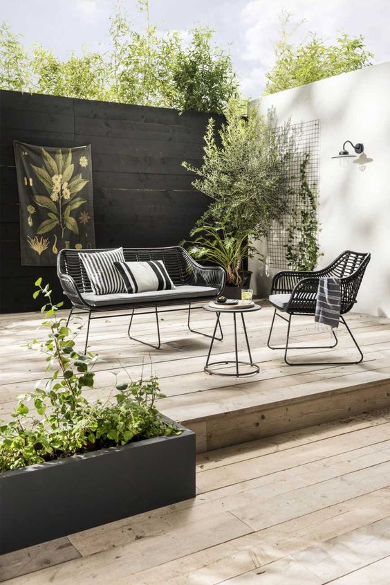 a modern Scandinavian terrace wiht a black fence, dark rattan furniture, greenery and trees is a lovely and cool outdoor space