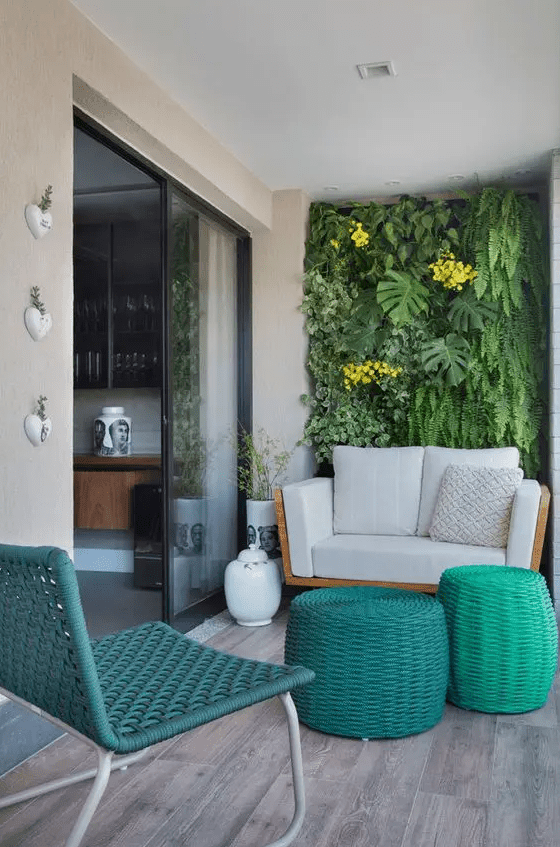 a modern and bright balcony with a living wall, a cozy loveseat, bright wicker side tables and a teal woven chair plus some blooms