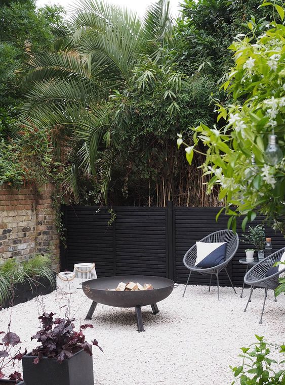 a modern and chic terrace with a black fence, modern furniture, a large fire pit, some greenery, candle lanterns around