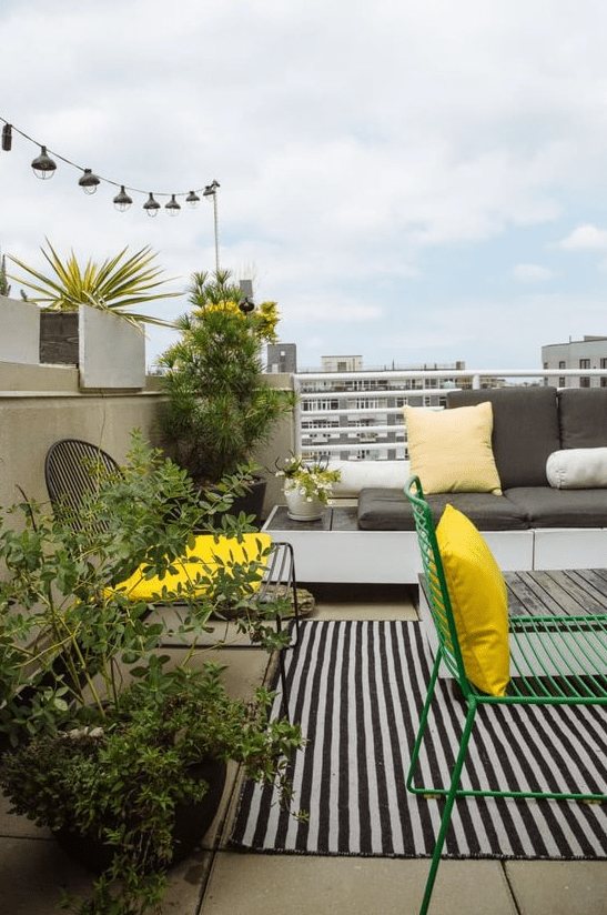 A modern balcony with a striped rug, a built in sofa with pillows, metal furniture, potted greenery and a city view