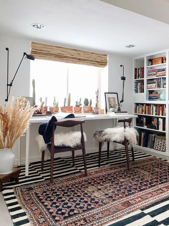 a modern boho home office with a built-in bookcase, a double desk, black chairs, layered rugs, a cacti collection and pampas grass