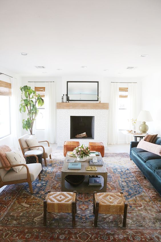 a modern boho living room with a fireplace, a navy sofa, a coffee table, neutral chairs, boho stools and some pretty decor