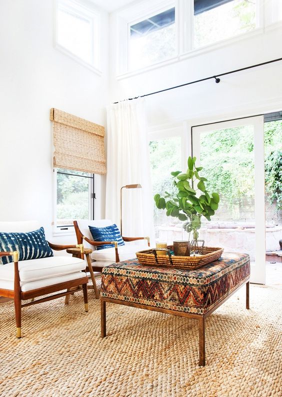 a modern boho living room with a jute rug, a bold boho ottoman, white chairs with coastal pillows, greenery and woven shades