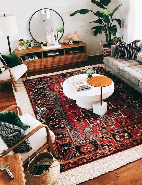 a modern boho living room with layered rugs, a grey sofa, white chairs, pillows, a sideboard, potted plants and candles